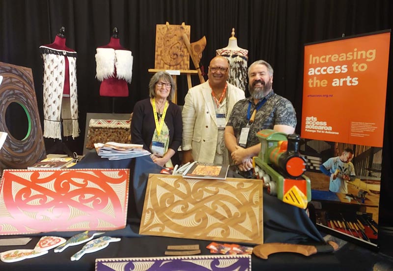 three people standing behind a display of carving and raranga  and in front of the Arts Access Aotearoa banner